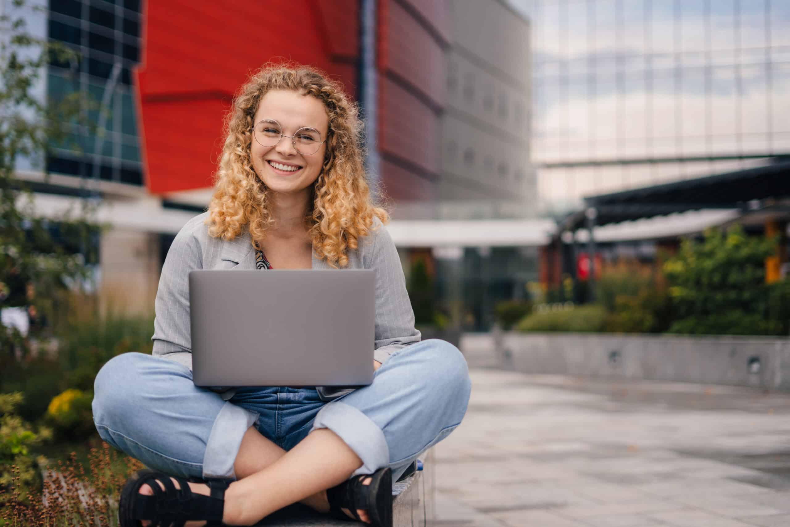 Étudiante souriante assise en tailleur avec son ordinateur portable, sur un campus