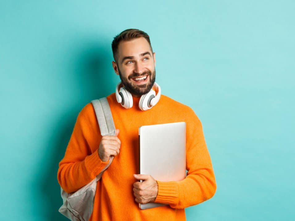 Un jeune homme souriant, sac à dos et ordinateur portable à la main
