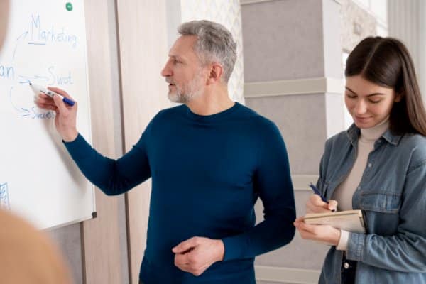 Un formateur explique un schéma au tableau à une apprenante qui prend des notes