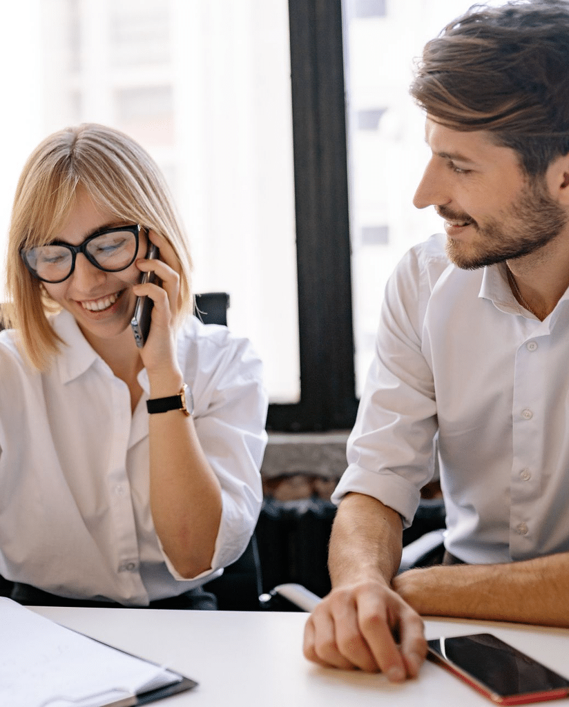 Femme au téléphone et homme souriant, assis côte à côte au bureau